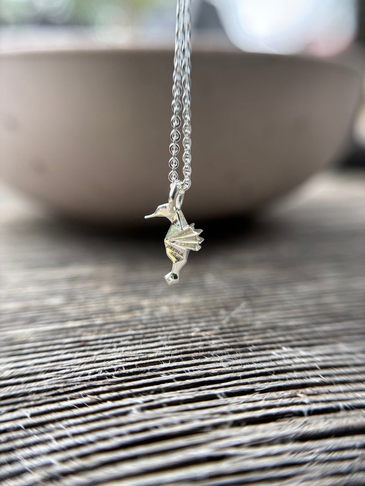 Collar de plata con un dije de caballito de mar estilo origami, visto de perfil sobre un fondo cn textura.