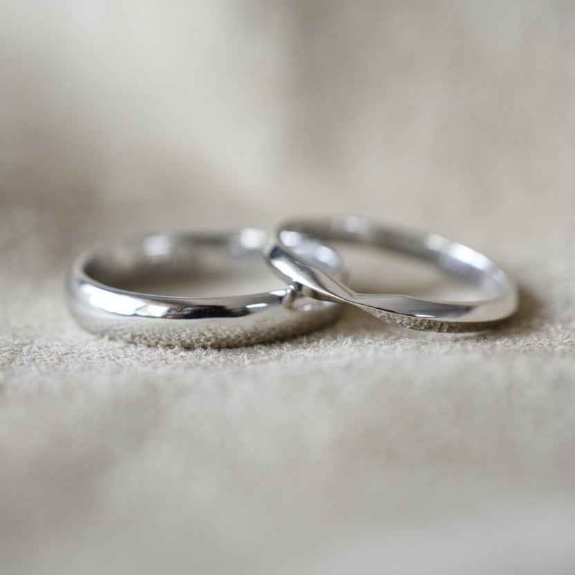 Vista detallada de dos argollas de matrimonio de oro blanco sobre tela suave. La argolla más ancha tiene perfil media caña y la más delgada perfil de cuchilla con un detalle de diseño central.