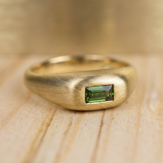Anillo chunky de oro amarillo con acabado satinado y un zafiro verde corte baguette engastado al ras, presentado sobre una superficie de madera clara.