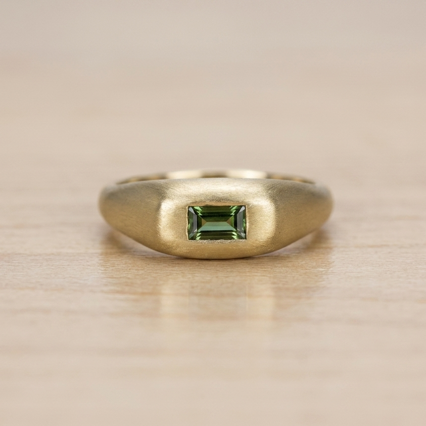Anillo chunky de oro amarillo con acabado satinado y un zafiro verde corte baguette engastado al ras, presentado sobre una superficie de madera clara.