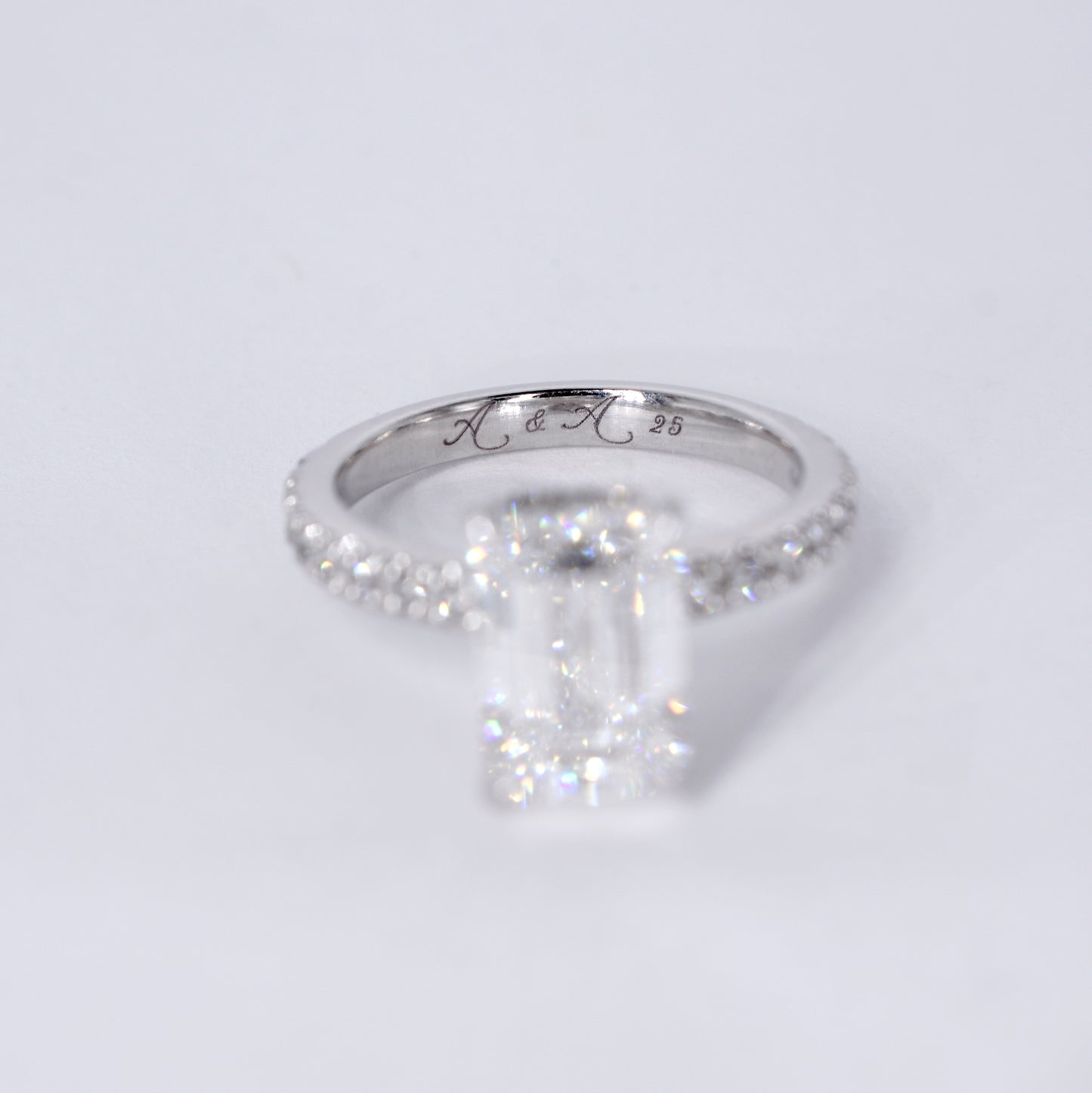 Anillo de compromiso de platino con un diamante central de corte esmeralda y una banda delgada con diamantes pequeños engastados en pavé. Vista frontal sobre un fondo blanco limpio y desenfocado.
