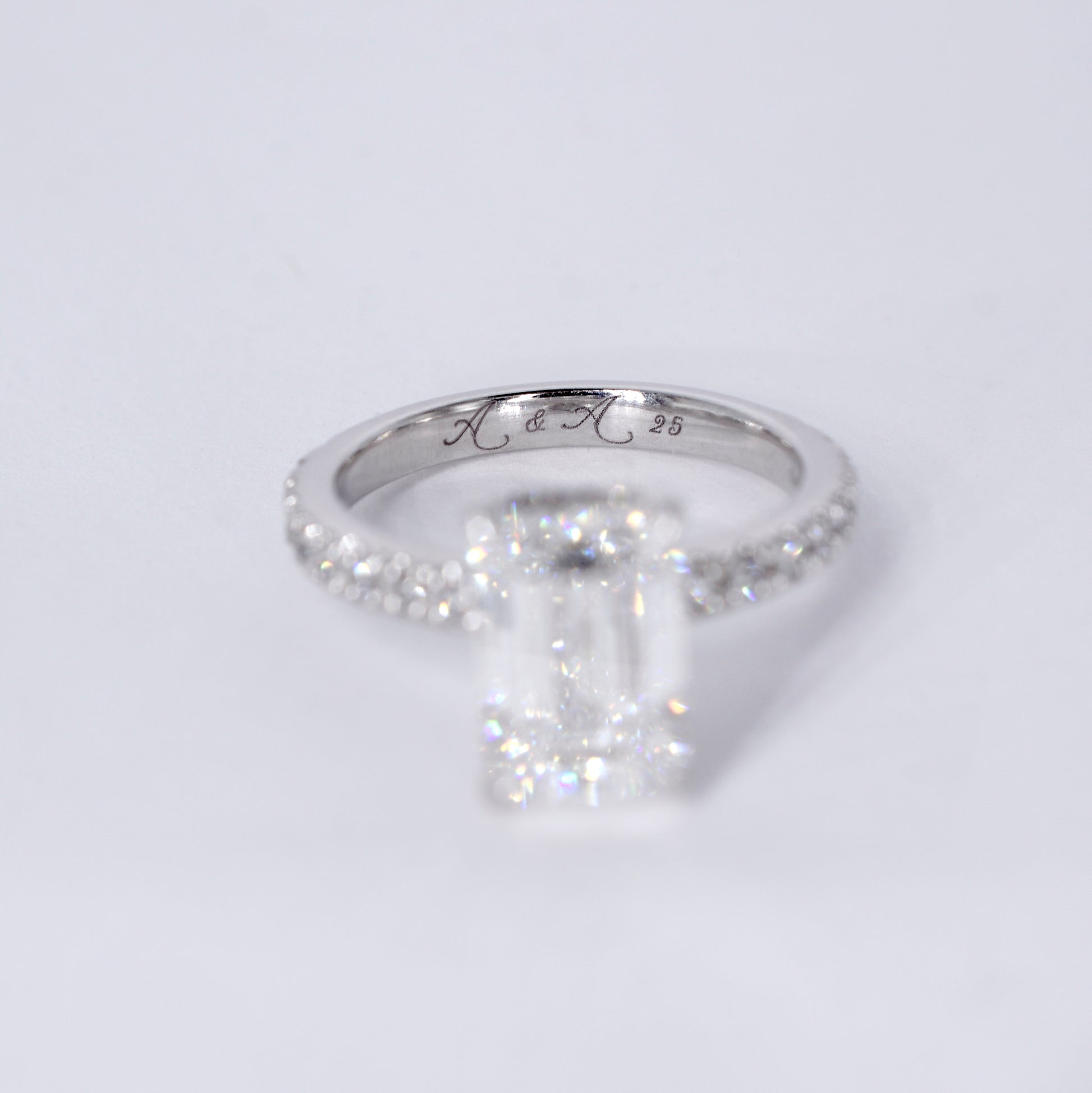 Anillo de compromiso de platino con un diamante central de corte esmeralda y una banda delgada con diamantes pequeños engastados en pavé. Vista frontal sobre un fondo blanco limpio y desenfocado.