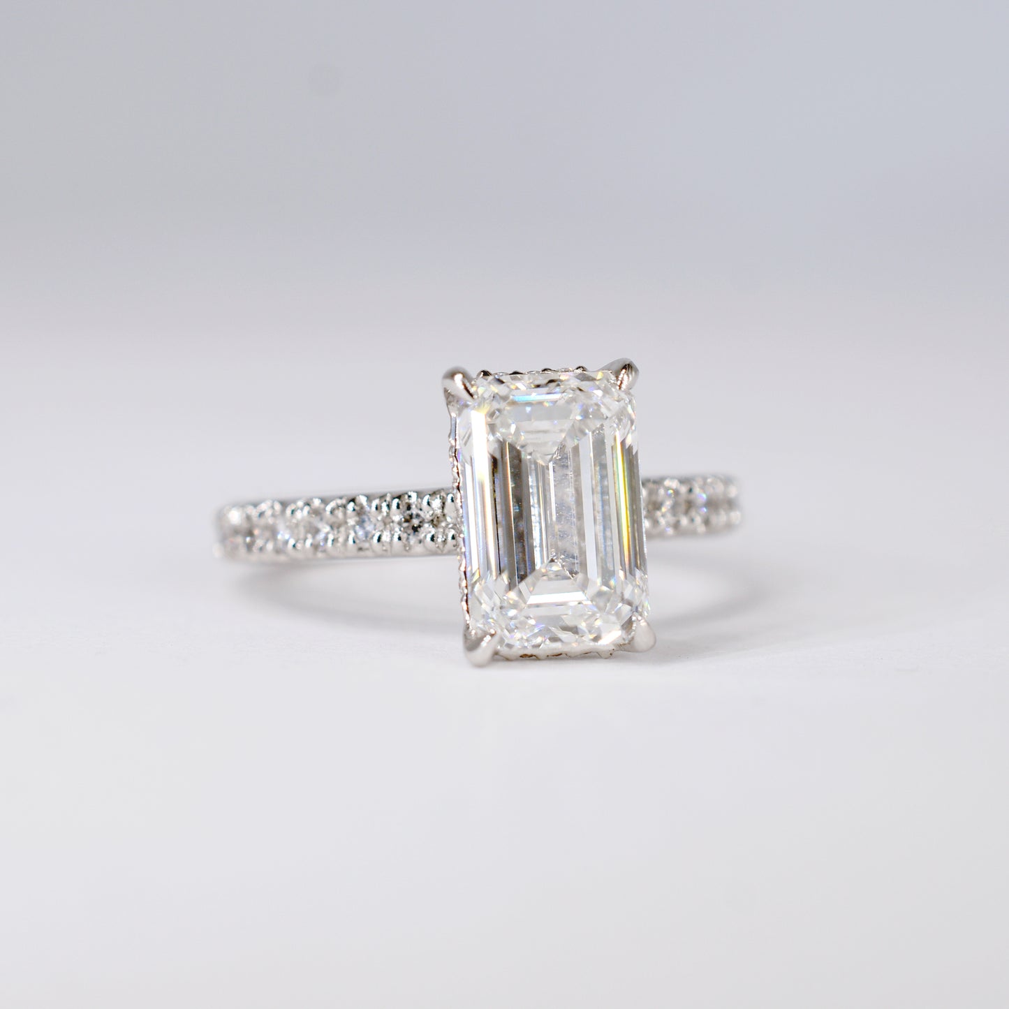 Anillo de compromiso de platino con un diamante central de corte esmeralda y una banda delgada con diamantes pequeños engastados en pavé. Vista frontal sobre un fondo blanco limpio y desenfocado.