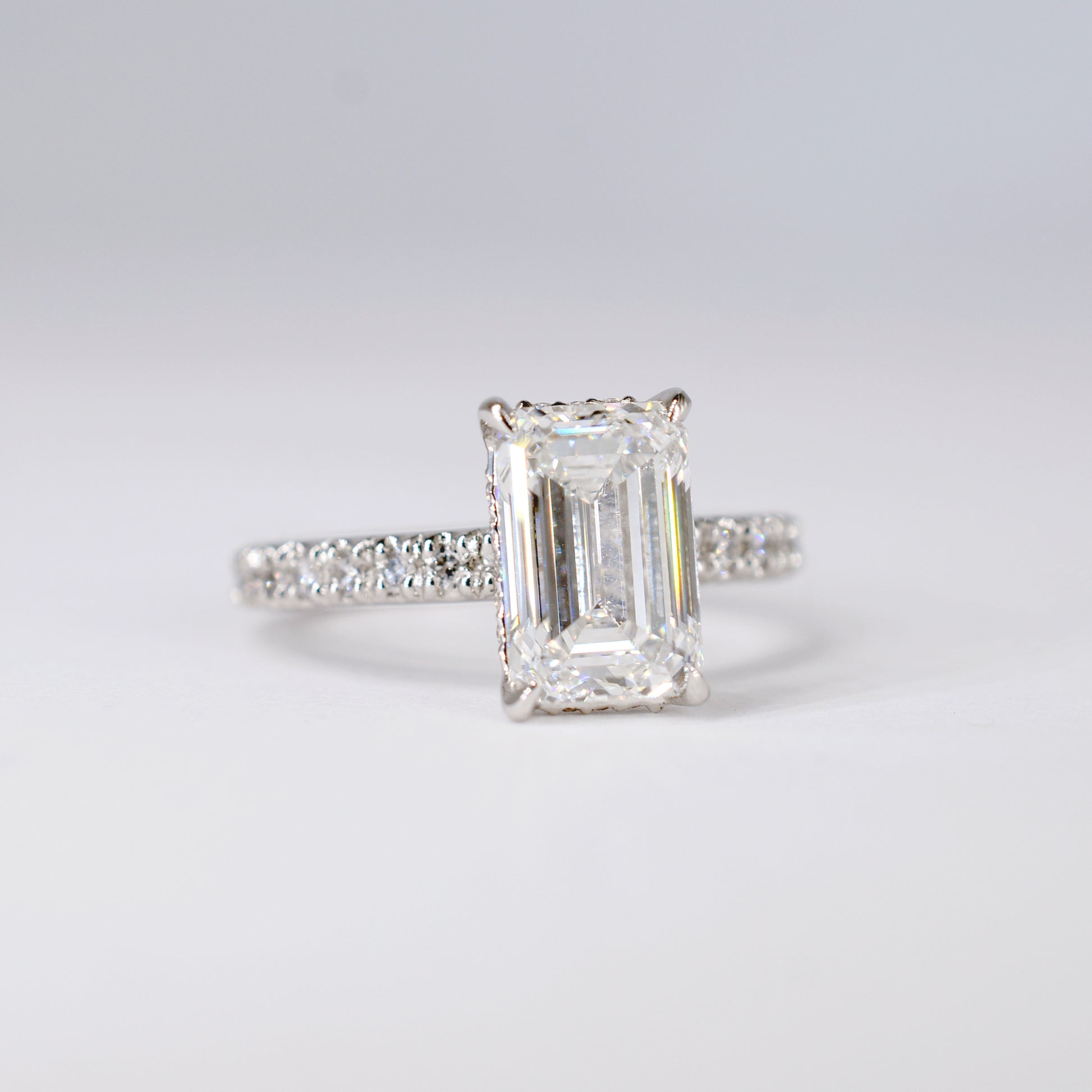 Anillo de compromiso de platino con un diamante central de corte esmeralda y una banda delgada con diamantes pequeños engastados en pavé. Vista frontal sobre un fondo blanco limpio y desenfocado.