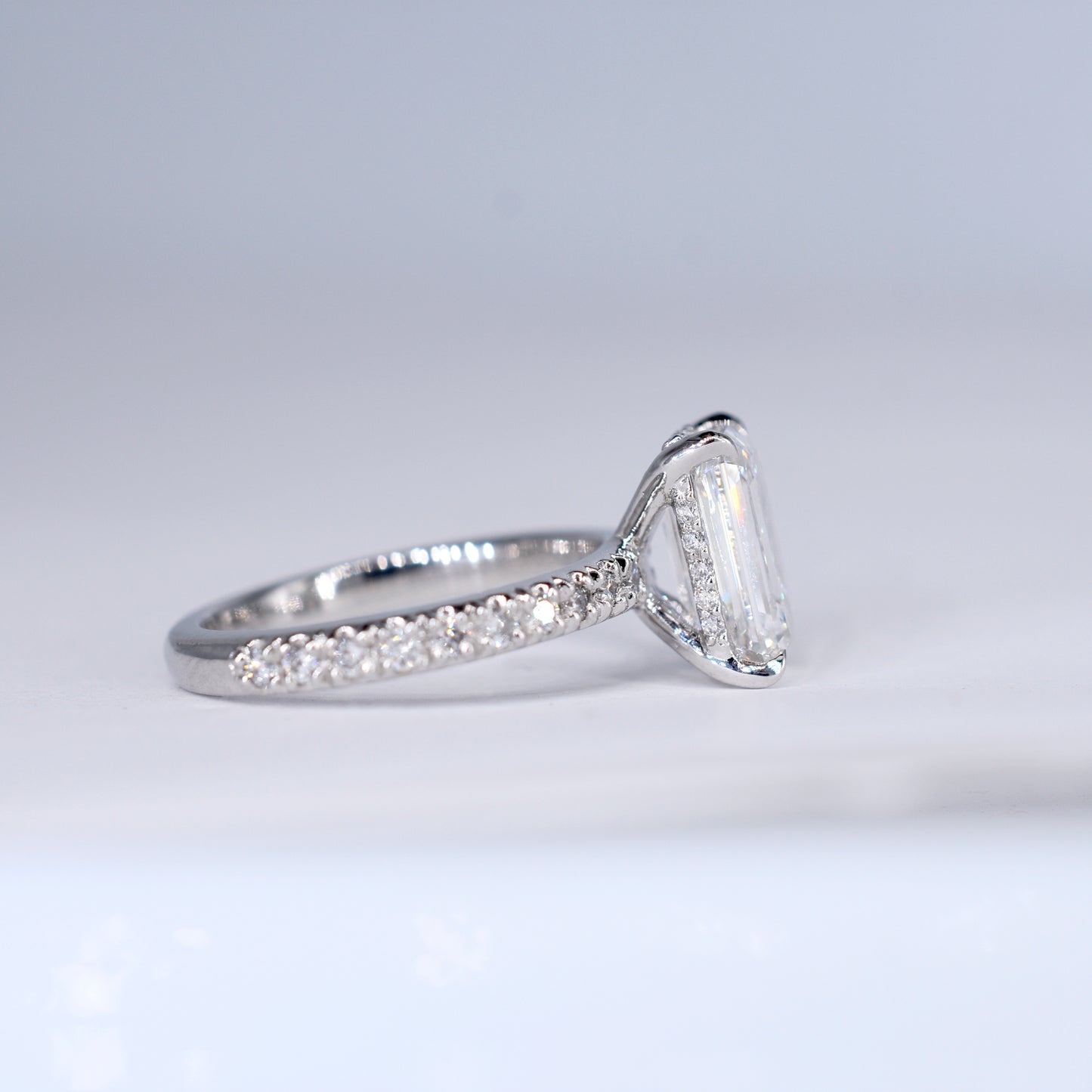 Anillo de compromiso de platino con un diamante central de corte esmeralda y una banda delgada con diamantes pequeños engastados en pavé. Vista frontal sobre un fondo blanco limpio y desenfocado.