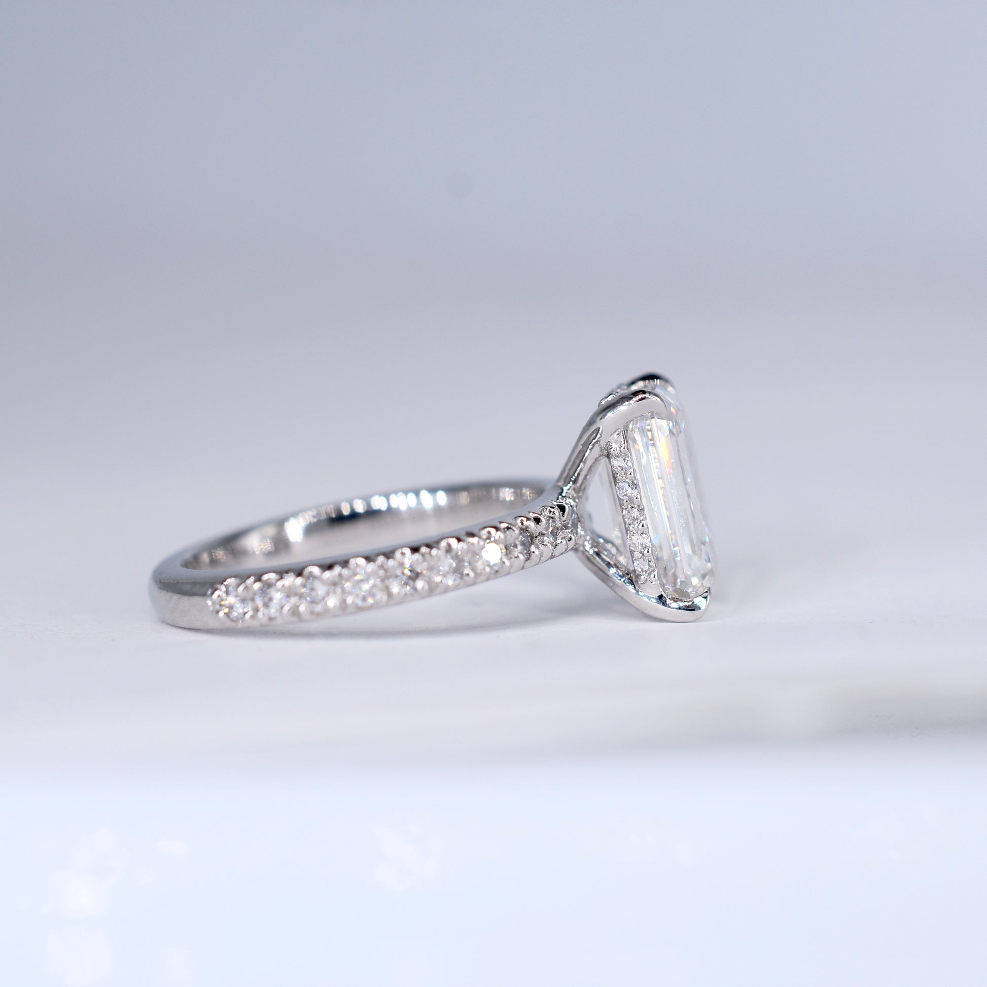 Anillo de compromiso de platino con un diamante central de corte esmeralda y una banda delgada con diamantes pequeños engastados en pavé. Vista frontal sobre un fondo blanco limpio y desenfocado.
