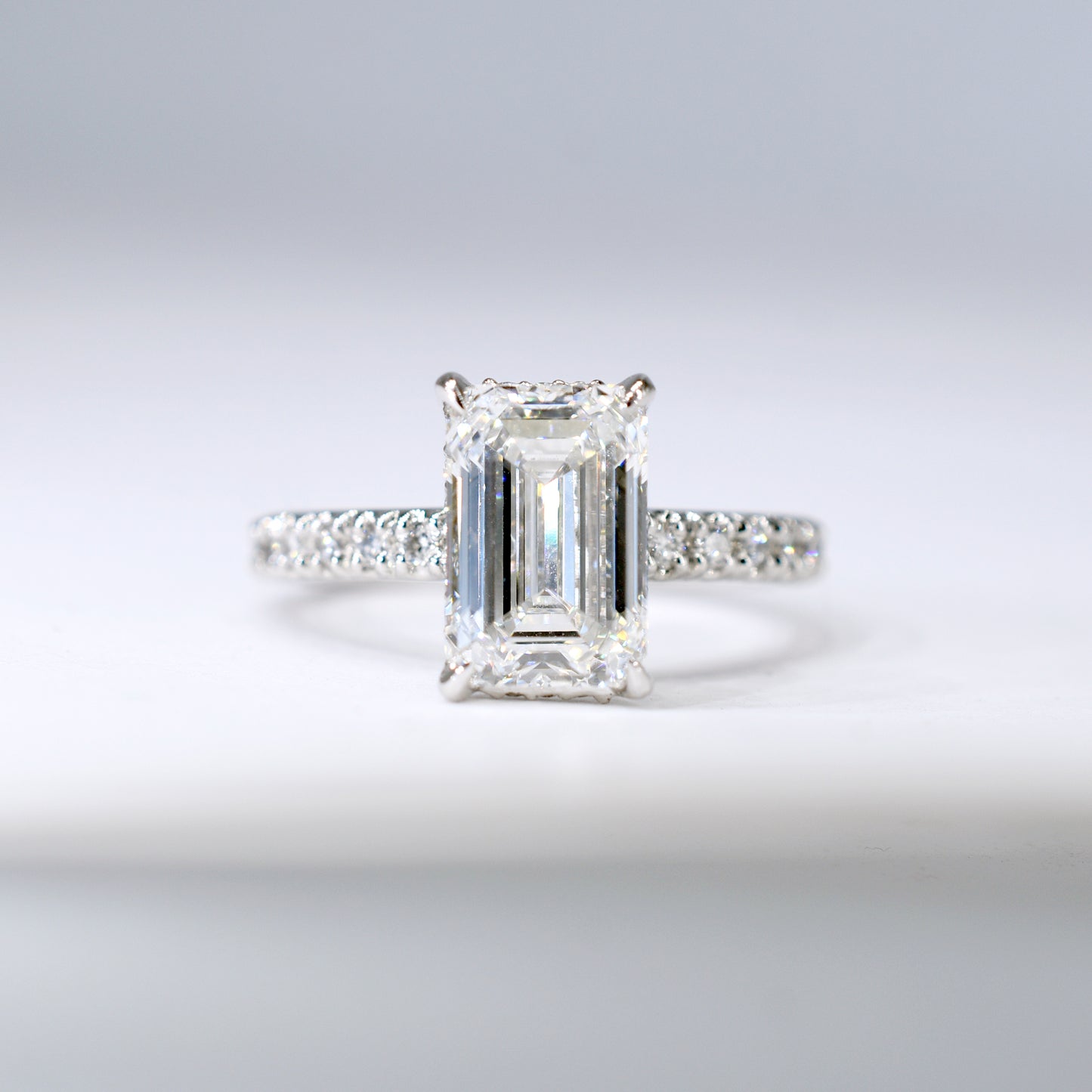 Anillo de compromiso de platino con un diamante central de corte esmeralda y una banda delgada con diamantes pequeños engastados en pavé. Vista frontal sobre un fondo blanco limpio y desenfocado.