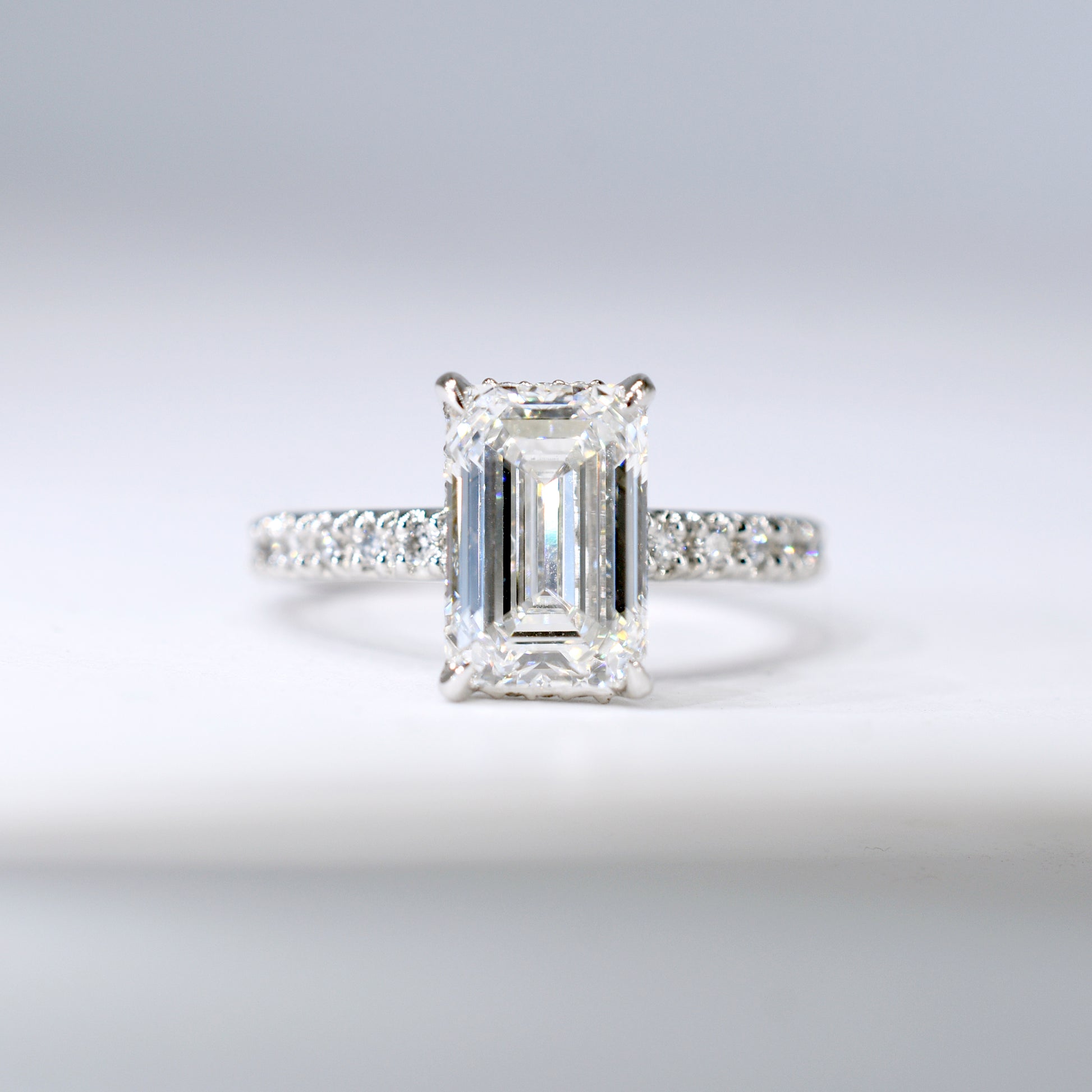 Anillo de compromiso de platino con un diamante central de corte esmeralda y una banda delgada con diamantes pequeños engastados en pavé. Vista frontal sobre un fondo blanco limpio y desenfocado.