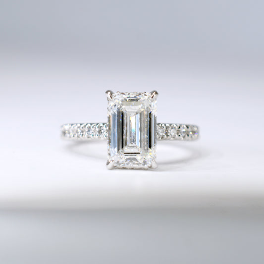 Anillo de compromiso de platino con un diamante central de corte esmeralda y una banda delgada con diamantes pequeños engastados en pavé. Vista frontal sobre un fondo blanco limpio y desenfocado.