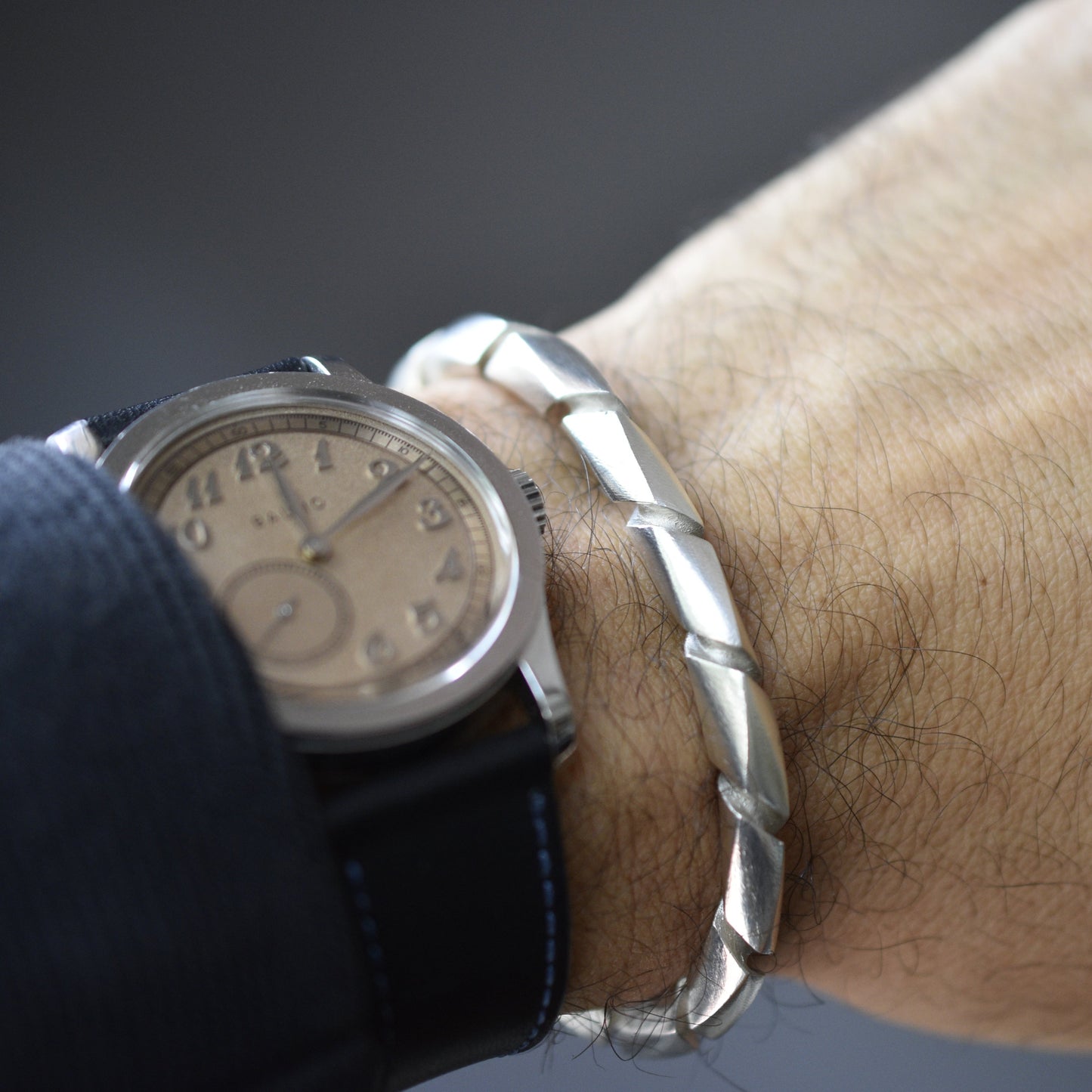 Primer plano de una muñeca masculina con un reloj de pulsera de esfera clara y una pulsera ancha de plata con un diseño trenzado o en espiral.