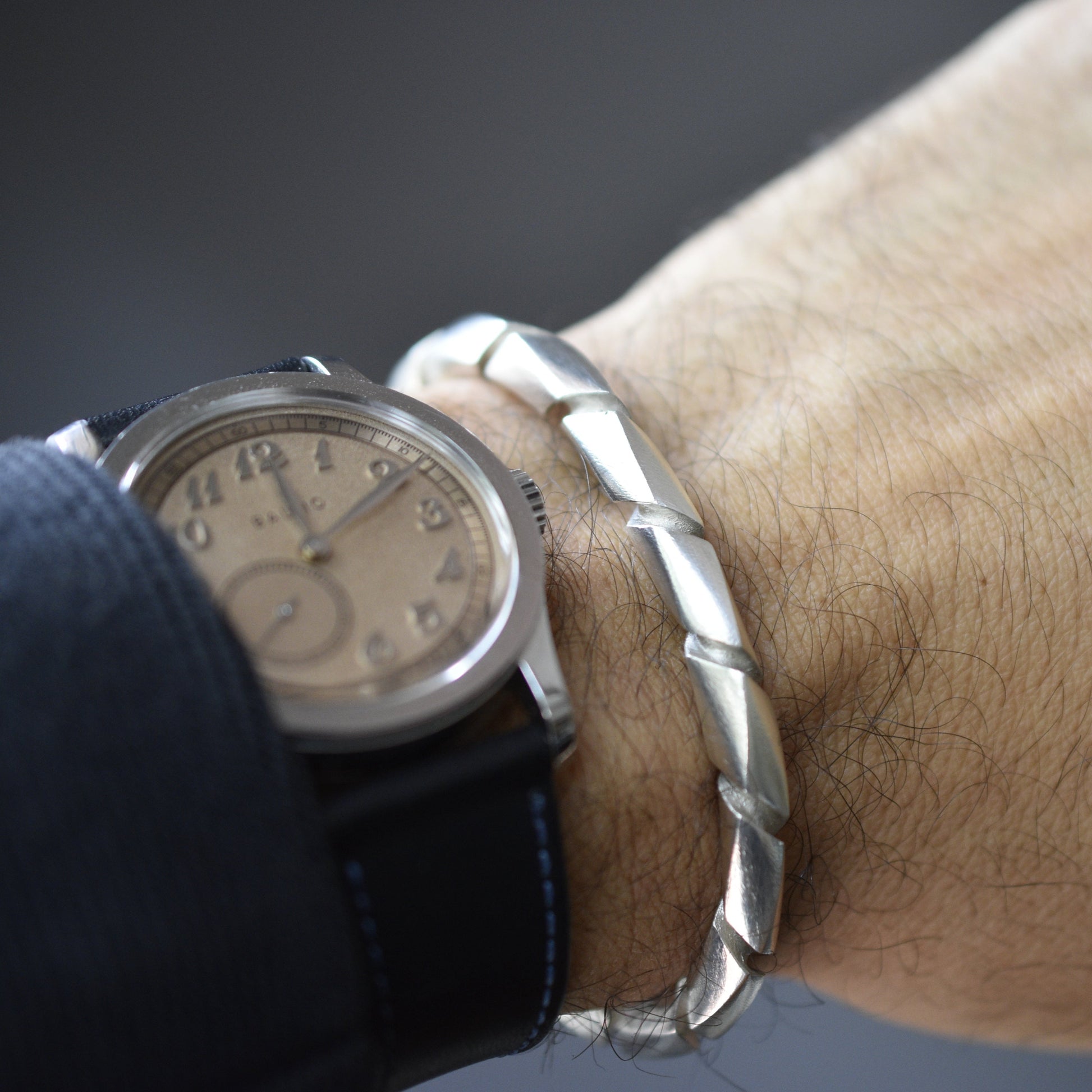 Primer plano de una muñeca masculina con un reloj de pulsera de esfera clara y una pulsera ancha de plata con un diseño trenzado o en espiral.